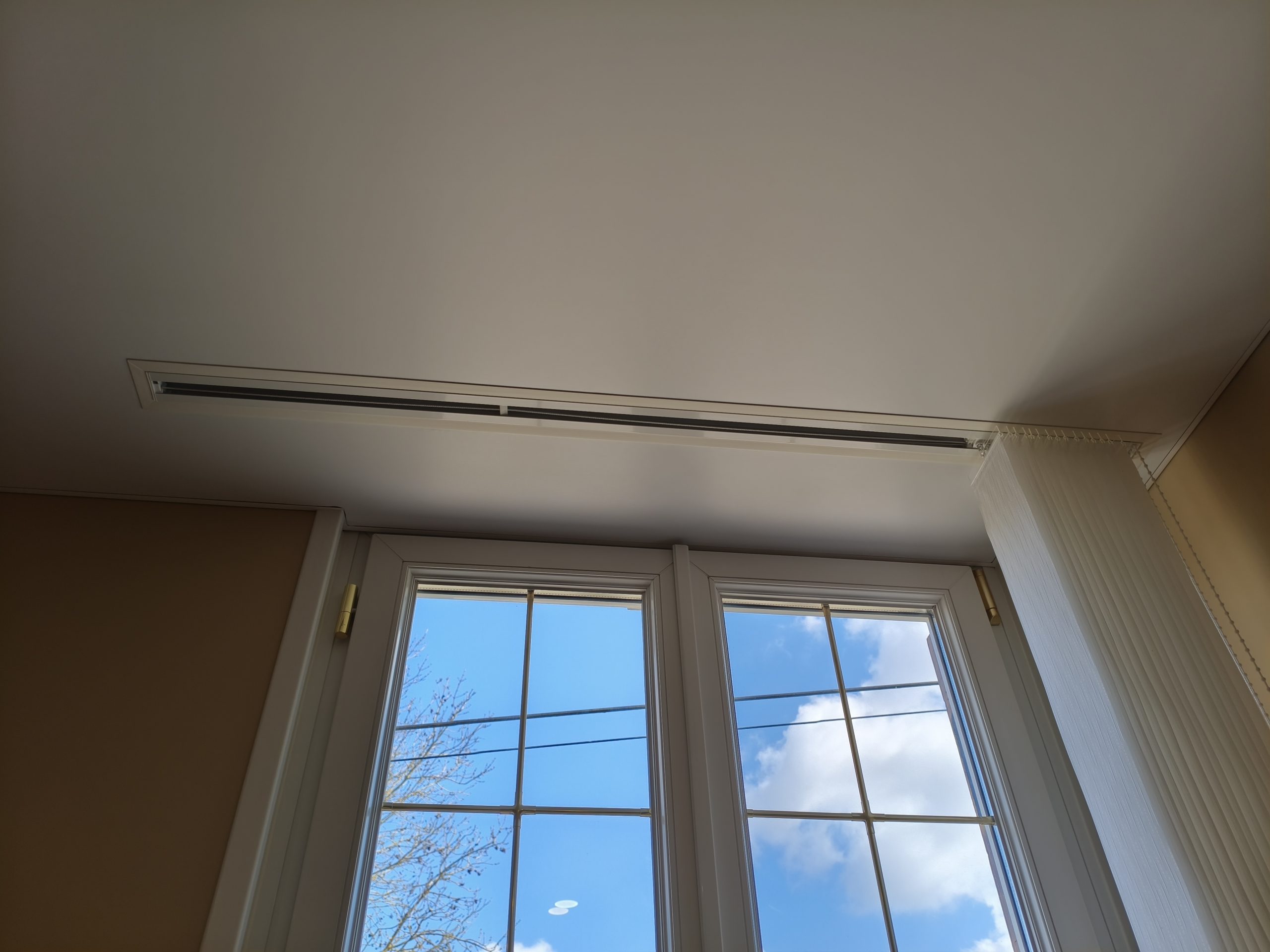 Détail du plafond tendu blanc au-dessus de la fenêtre et du rail du store à bandes verticales.